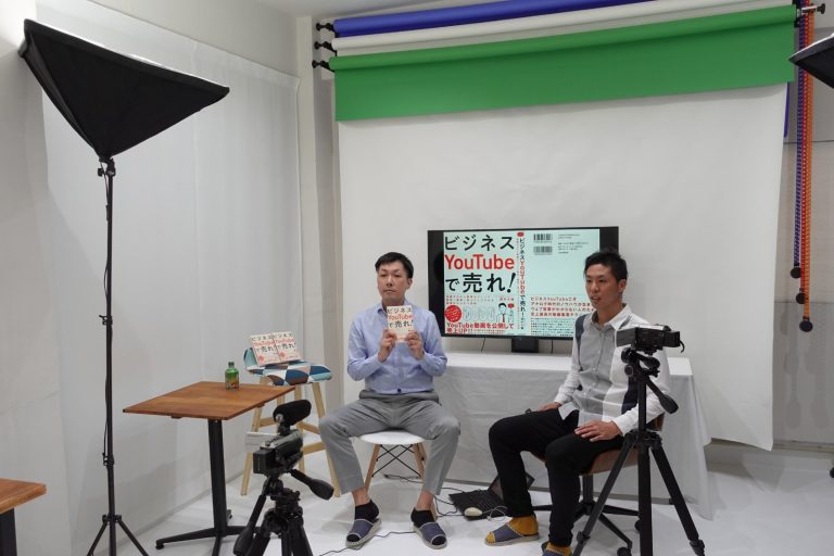 白ホリスタジオでのインタビュー収録の風景。50型モニターの前で2人が対談し、三脚のカメラ2台とソフトボックスで撮影している